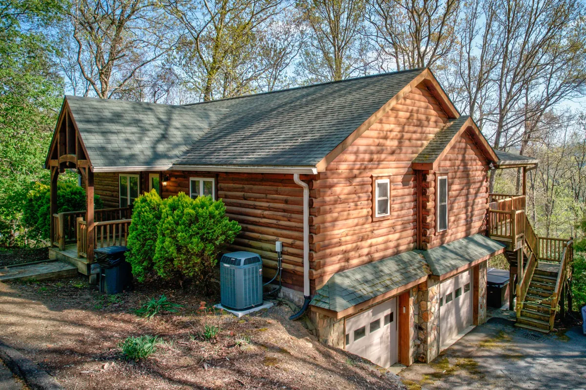 A cozy log cabin surrounded by lush greenery, with stairs leading up to an inviting upper deck. The setting suggests space for family gatherings, outdoor barbecues, and enjoying the fresh mountain air.