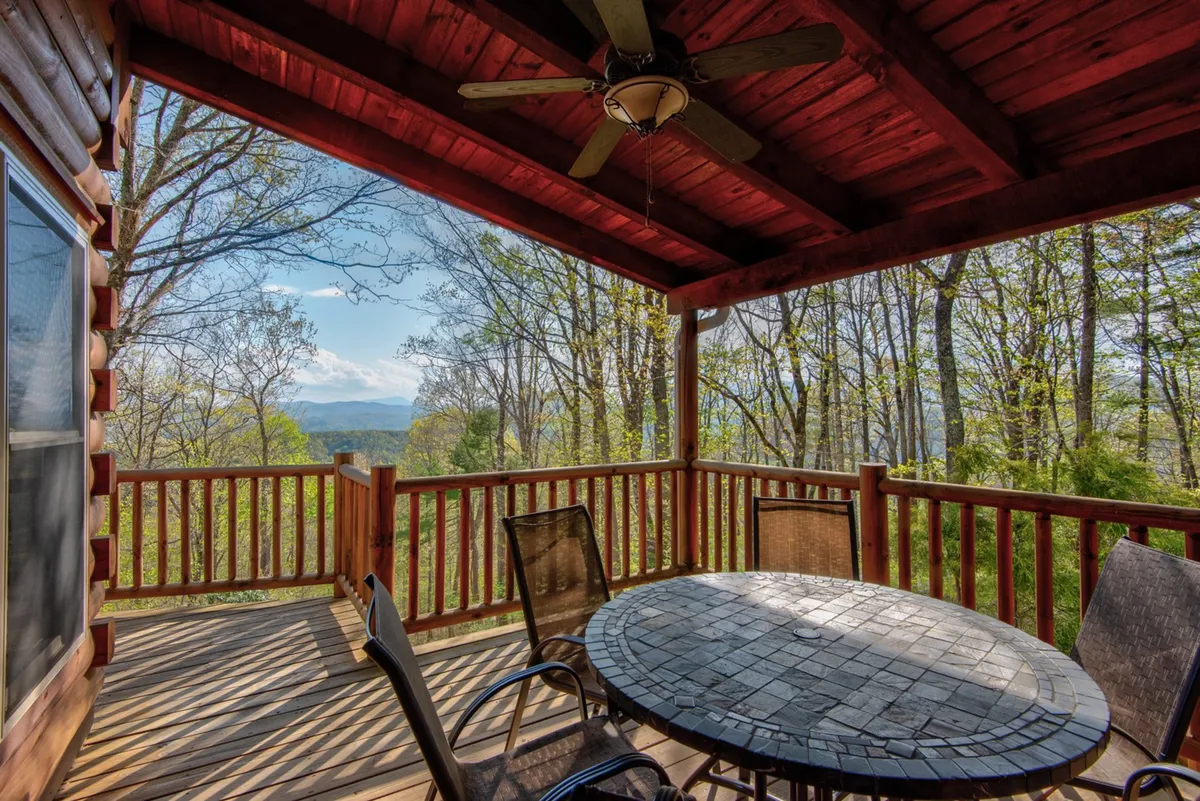 A spacious wooden deck is outfitted with a round dining table and chairs, all oriented toward a mountain view framed by surrounding trees. The scene suggests a perfect spot for group gatherings, outdoor meals, or quiet moments amid nature.