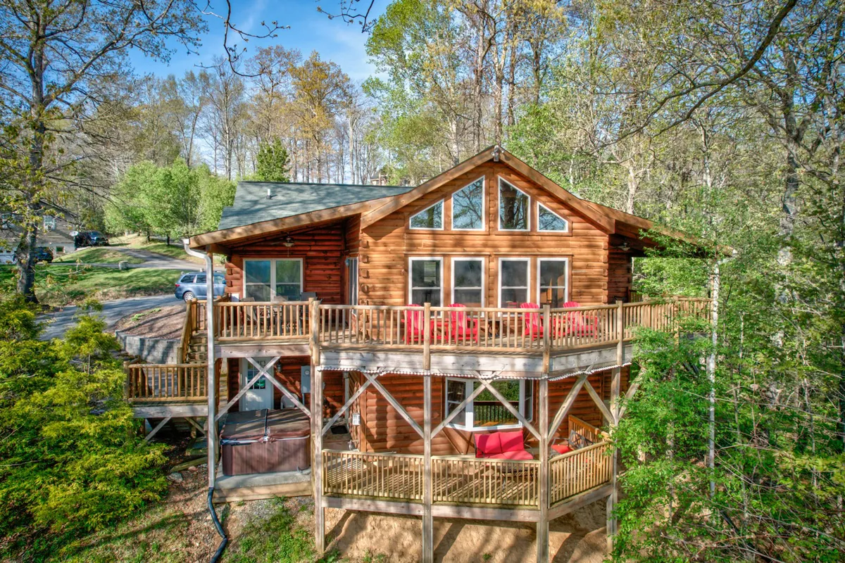 The image showcases the log cabin's inviting exterior, highlighting expansive windows that flood the interior with natural light, a spacious wooden deck with vibrant red outdoor furniture, and sweeping scenic views—perfect for gathering with family or friends. The surrounding mountain landscape creates a tranquil, nature-filled backdrop, while the deck is set up for sunset dinners or peaceful morning coffee.