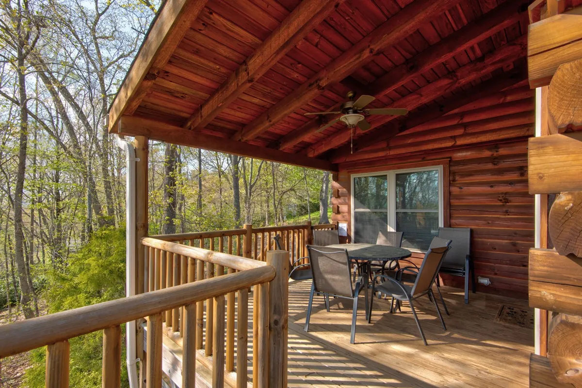 This exterior shot captures a wooden deck with a cozy outdoor dining set, log cabin wall, and surrounding trees. The deck’s railing and furniture suggest a welcoming space for outdoor dining or sunset toasts, while the treetops hint at a secluded, nature-rich location. The mood is tranquil and perfect for intimate moments.