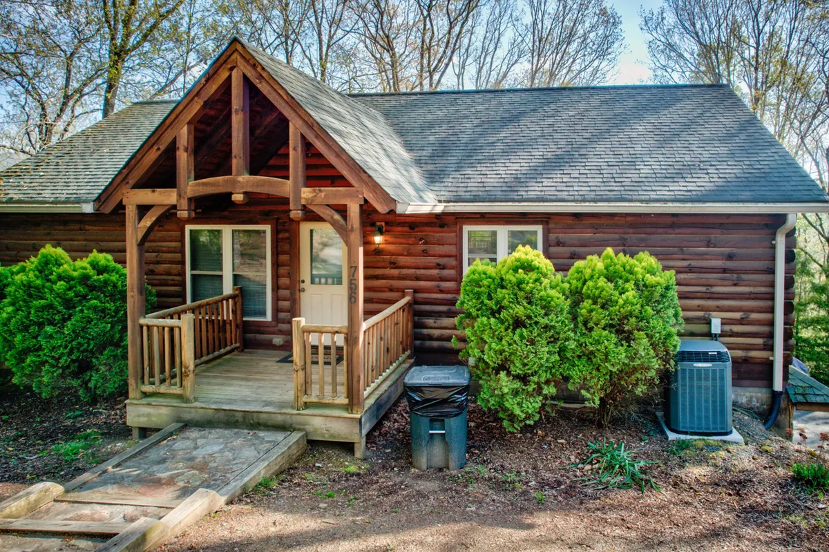 A welcoming wooden porch at the front of the cabin, featuring a sitting area surrounded by greenery. The roof structure and bushes frame the entrance, evoking a sense of arrival and the natural, tranquil mountain environment.
