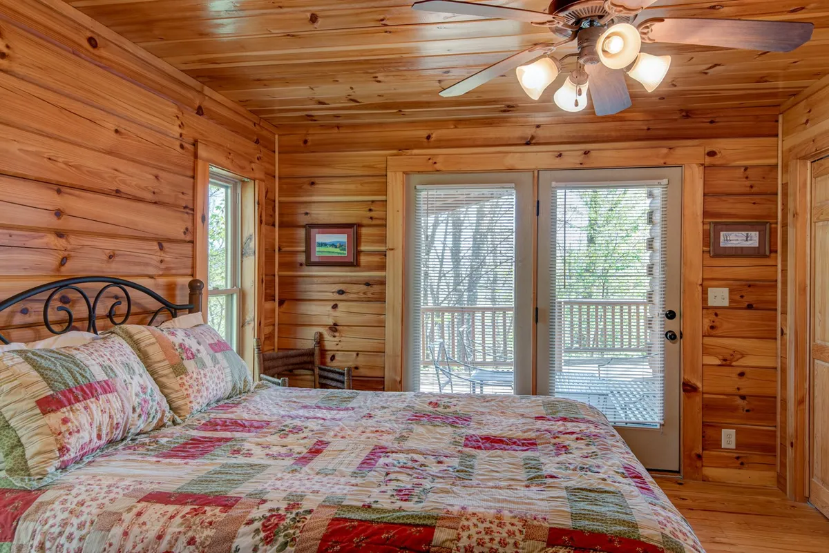 A cozy bedroom with a quilted bed, wooden wall paneling, and double doors that open directly onto a deck. The room is bathed in natural light, offering both comfort and immediate access to an outdoor retreat, emphasizing privacy and serenity.