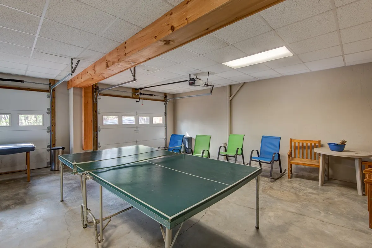 A lighthearted inside scene showing a spacious area with a ping pong table, colorful chairs, a wooden bench, and a small round table. The space is arranged for casual gatherings, tournaments, and socializing, capturing the spirit of group activities.