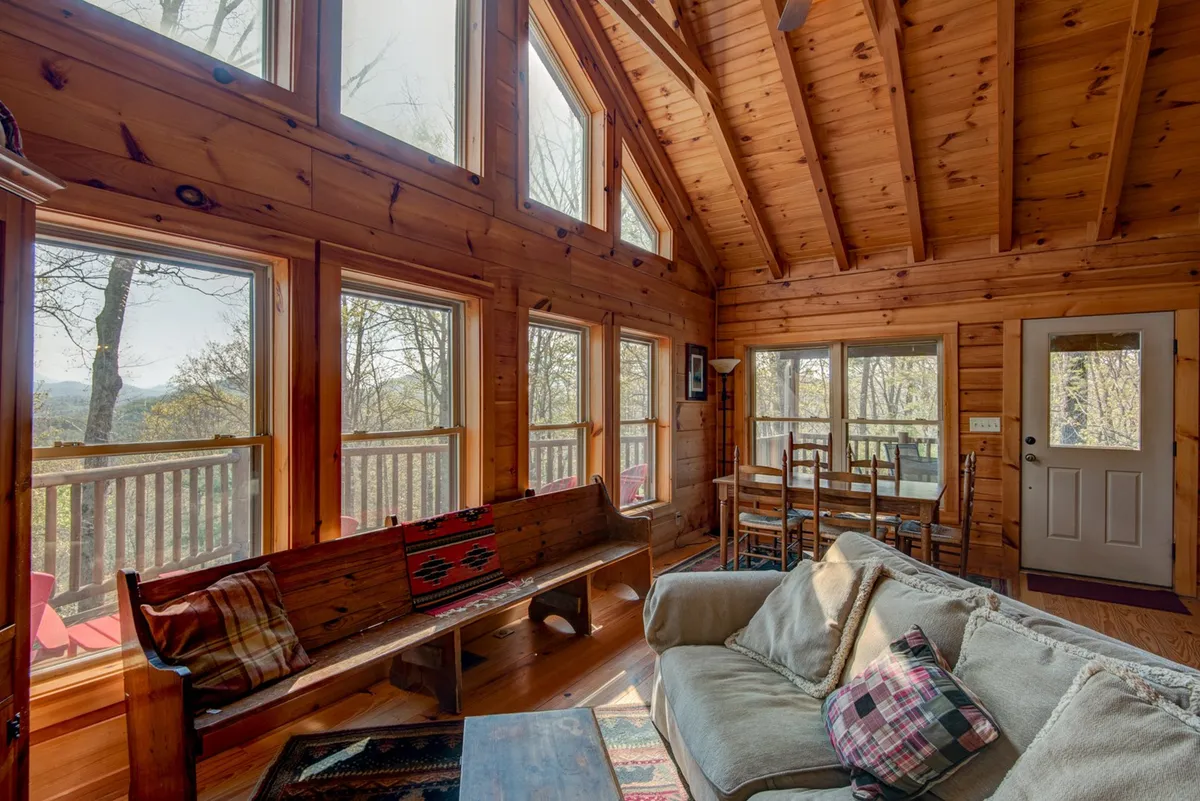 A warm, open-concept interior with exposed wooden beams, a cozy sofa, a dining table for family meals, and large windows showcasing abundant natural light and outdoor views. The arrangement highlights seamless flow between living and dining spaces.