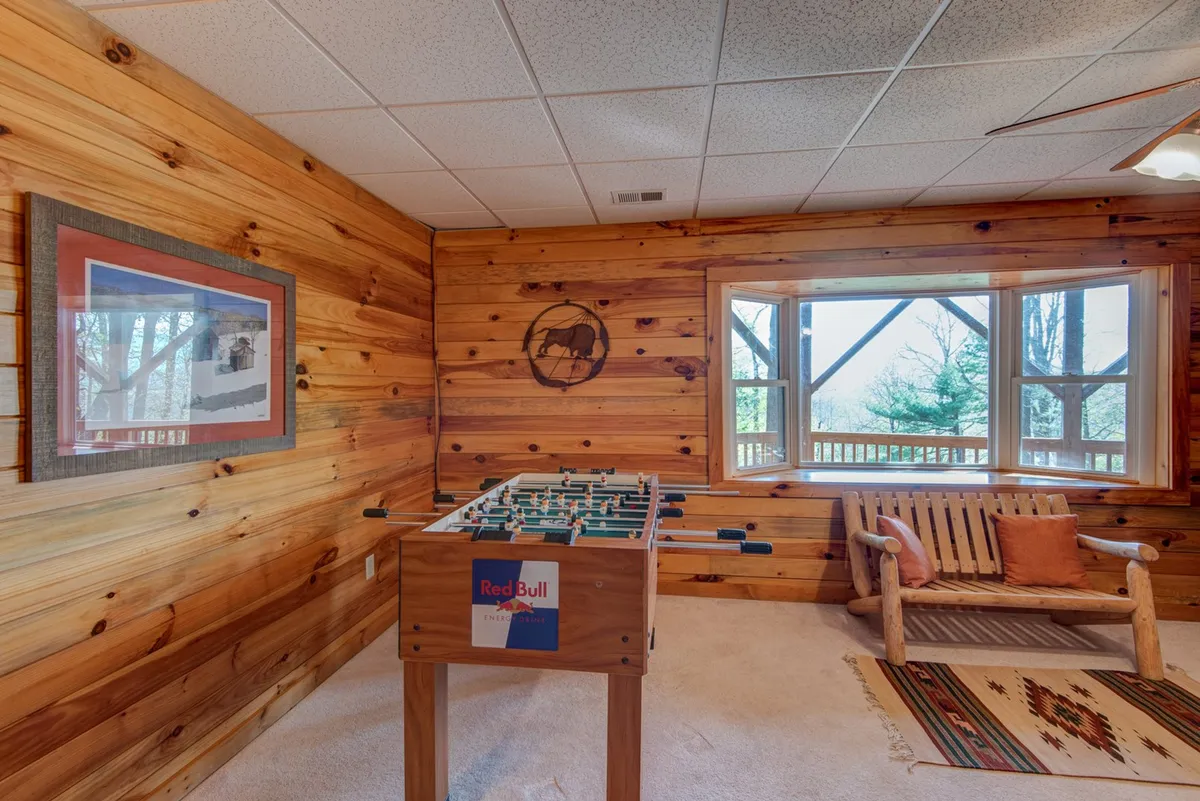This image captures a playful, inviting interior space centered around a foosball table, with large windows letting in natural light. The room includes a wooden bench and decorative wall art, and groups are seen socializing and enjoying friendly competitions.